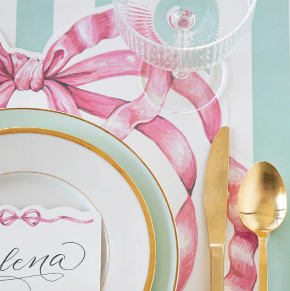 Elegant table setting with gold-rimmed plates, gold flatware, crystal glass, and a Hester & Cook Die-cut Pink Bow Placemat (12 Sheets) from the Pink Bow Collection. A cursive name card rests gracefully on the plate.