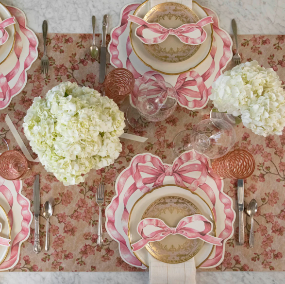 A table set for four with gold-trimmed plates, pink and white napkins tied in bows, Hester & Cook Die-cut Pink Bow Placemat (12 Sheets), pink glasses, silver cutlery, and white hydrangea centerpieces on a floral runner.
