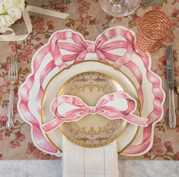 A table setting features a Hester & Cook Die-cut Pink Bow Placemat, scalloped and small plates with pink bows, neatly arranged silverware, a pink glass, and a white napkin.