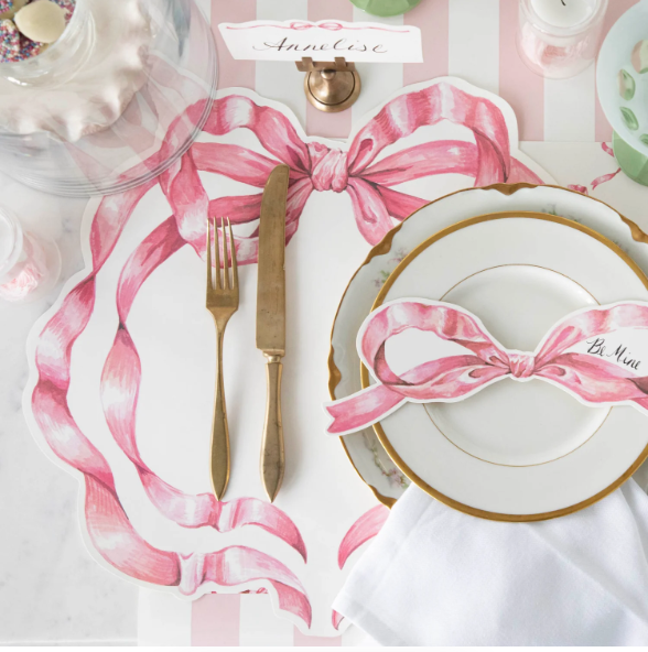 A place setting with gold-rimmed plates, gold cutlery, a white napkin, and the Hester & Cook Die-cut Pink Bow Placemat featuring a pink ribbon design. A handwritten name card for Annelise completes the Pink Bow Collection look.