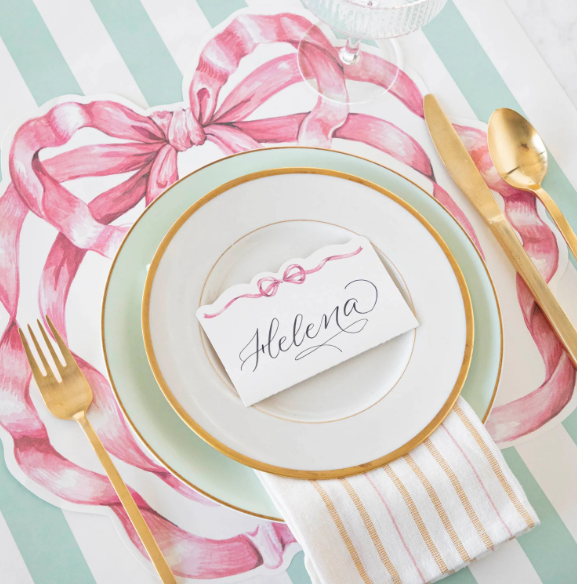 A table setting with gold cutlery, a white plate with gold trim, a striped napkin, and a "Helena" place card sits atop Hester & Cook's Die-cut Pink Bow Placemat. Mint green stripes and pink bow motifs complete the look.