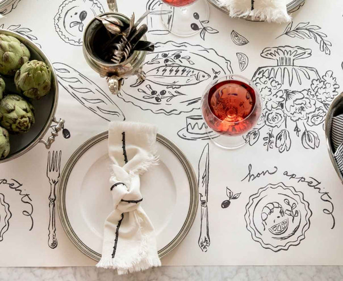 A table set with a white plate, napkin, cutlery, glass of rosé, artichokes in a bowl, and the Hester & Cook Dinner Party Runner (20x25"), featuring black food and floral sketches.