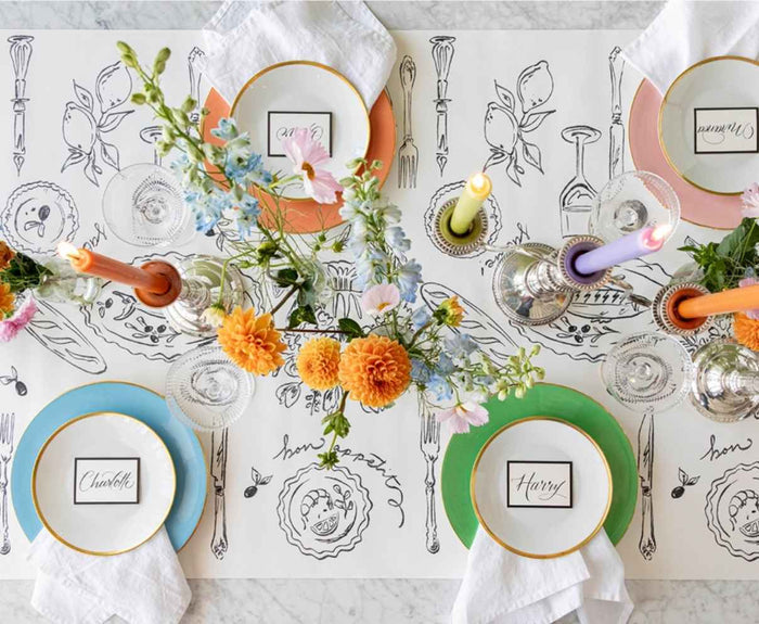 Bright spring table with orange, green, and peach plates, white napkins, name cards, yellow-orange flowers, pastel candles, glassware, and a Hester & Cook Dinner Party Runner (20x25") in black-and-white illustration.