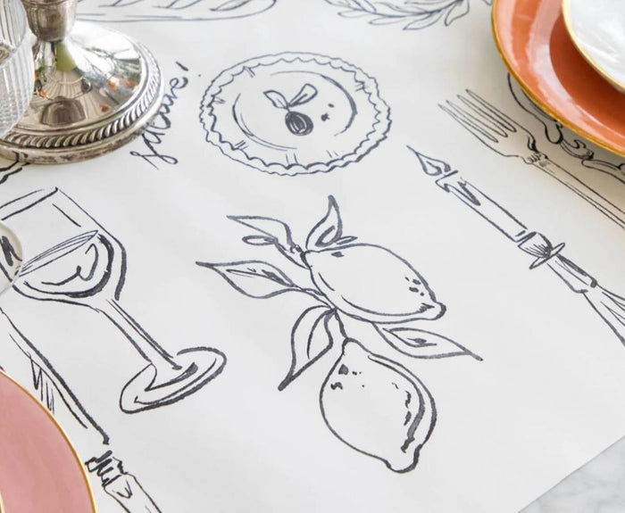 A tabletop features the Hester & Cook Dinner Party Runner - 20x25", a white paper runner with black lemon line drawings, accented by pink-rimmed plates, a wine glass, a fork, and a silver candlestick.