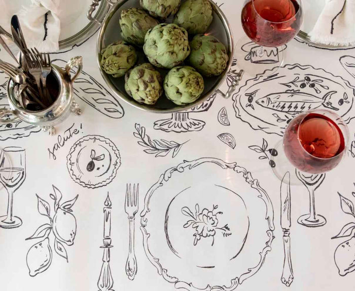 A table set with hand-drawn black and white placemats, artichokes, red wine glasses, and utensils paired with the Hester & Cook Dinner Party Runner (20x25"), featuring sketches of plates, cutlery, wine glasses, lemons, and a fish.