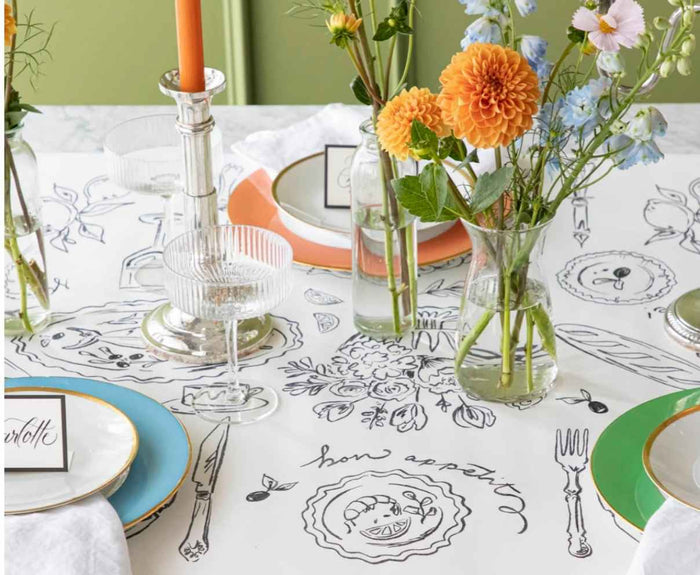 A table set for a meal with colorful plates, glassware, orange candles, and bright flowers on a Hester & Cook Dinner Party Runner (20x25"), featuring whimsical black line drawings and "bon appétit.