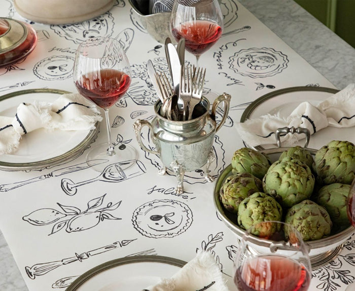 A table set with plates, silverware, napkins, wine glasses of rosé, artichokes, and a utensil holder sits atop the Hester & Cook Dinner Party Runner (20x25"), featuring black line art and food illustrations.