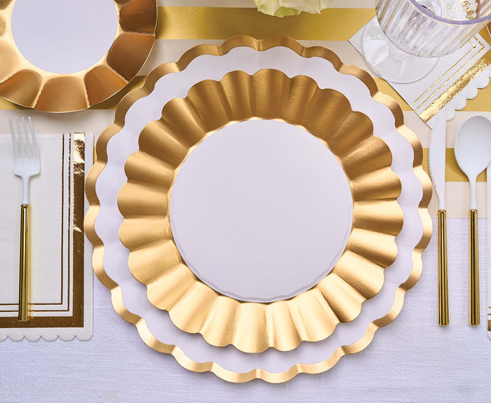 A table setting with Sophistiplate’s Dinner Plate Gold & White/8 pkg, gold-accented cutlery, a napkin, and a clear cup arranged on a white and gold striped tablecloth.