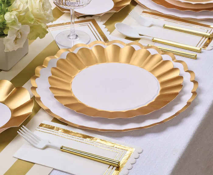 A close-up of a table set with Sophistiplate Dinner Plate Gold & White/8 pkg, gold-accented cutlery, a white napkin with gold trim, and a centerpiece of white flowers on a white tablecloth.