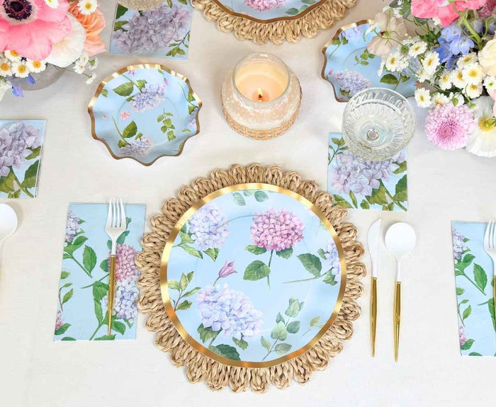 A table setting with Sophistiplate's Dinner Plate Hydrangea - 8pk, gold-rimmed chargers, gold and white cutlery, floral napkins, crystal glassware, a lit candle, and bouquets of pink, white, and purple flowers.