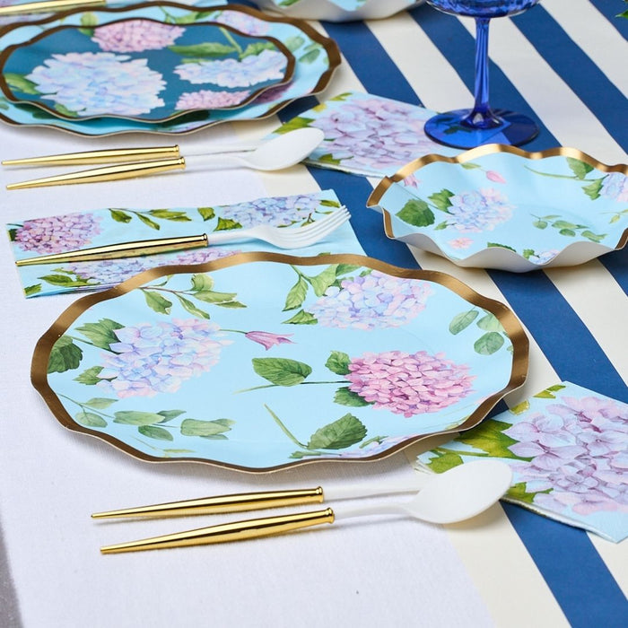 A close-up of a table set with Sophistiplate Dinner Plate Hydrangea - 8pk, showcasing blue floral plates with pink and purple hydrangeas, gold and white cutlery, a blue and white striped tablecloth, and a blue glass.