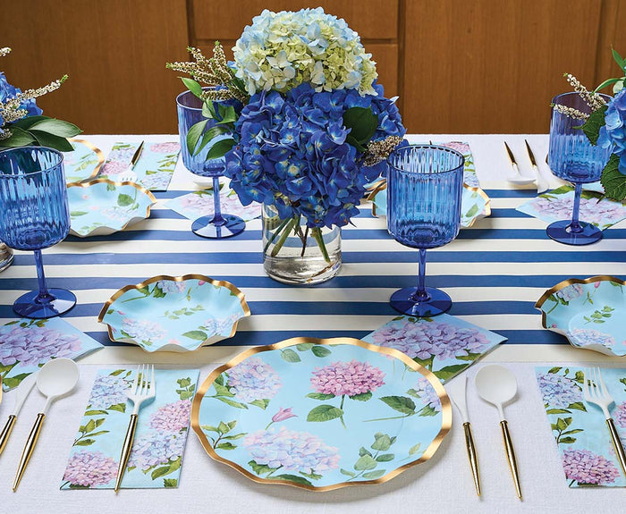A meal table features Sophistiplate Dinner Plate Hydrangea (8pk), blue hydrangea centerpieces, blue glassware, floral napkins, white utensils, and a blue-and-white striped table runner.