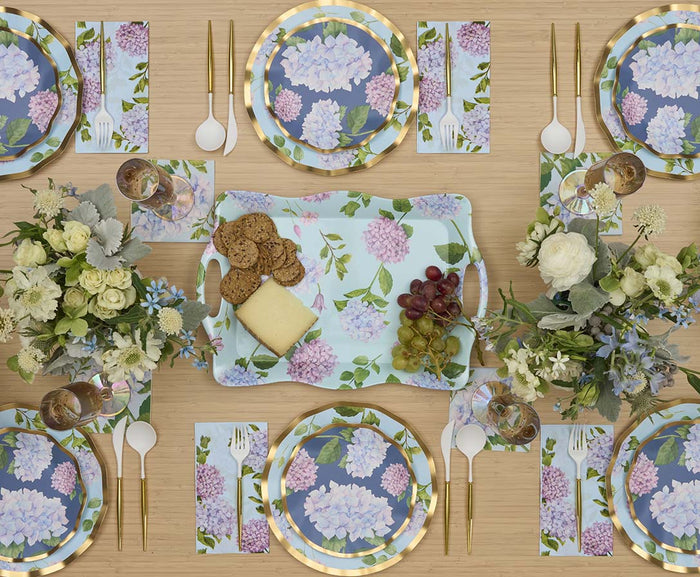 A table set for six features Sophistiplate Salad Plate Hydrangea (8pk) with gold utensils, wine glasses, and napkins on light wood. A centerpiece tray with cheese, crackers, and grapes is surrounded by white and green floral arrangements.