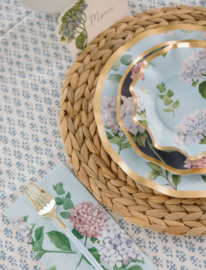 A table setting with Sophistiplate's Hydrangea Foil appetizer/dessert bowls (8pk), a woven placemat, floral napkin, gold fork and knife, and a "Marci" name card on a patterned tablecloth.