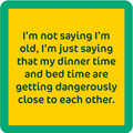 The Drinks On Me Dinner Time Coaster features a yellow square with a green border and the text: "I’m not saying I’m old, I’m just saying my dinner time and bed time are getting dangerously close"—a perfect quirky gift or funny drink coaster.