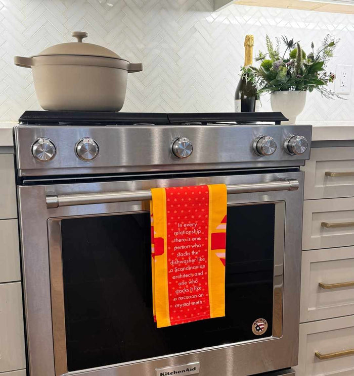 Add humor to your kitchen with the Drinks On Me Dishwasher Dish Towel, shown hanging on a modern stove beside a beige pot, champagne bottle, and floral arrangement—great as a unique housewarming gift or kitchen decor.