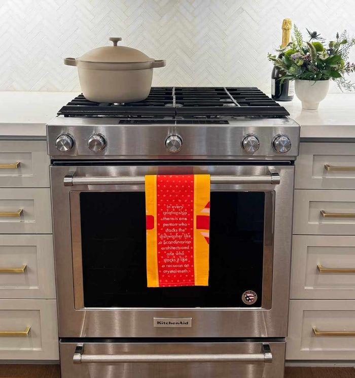 A modern stainless steel stove features a beige pot on the burner and a Drinks On Me Dishwasher Dish Towel hanging on the oven door. White cabinets, flowers, and a champagne bottle decorate the counter in the background.