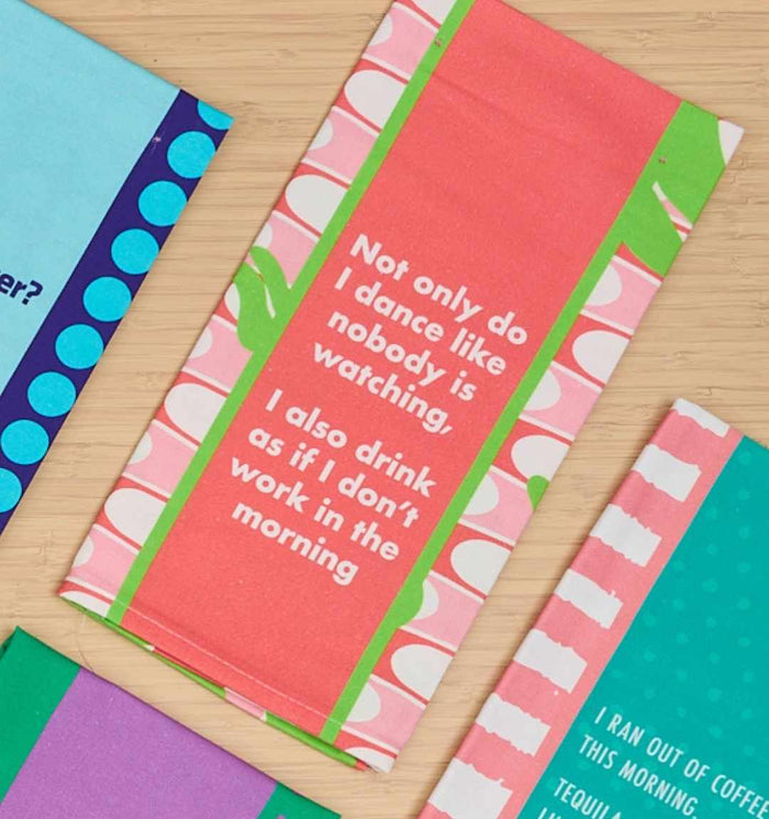 A pink and green “Don’t work in Morning” dish towel by Drinks On Me says, “Not only do I dance like nobody is watching, I also drink as if it doesn’t work in the morning”—a humorous housewarming gift. Other colorful cards are nearby on a wooden surface.