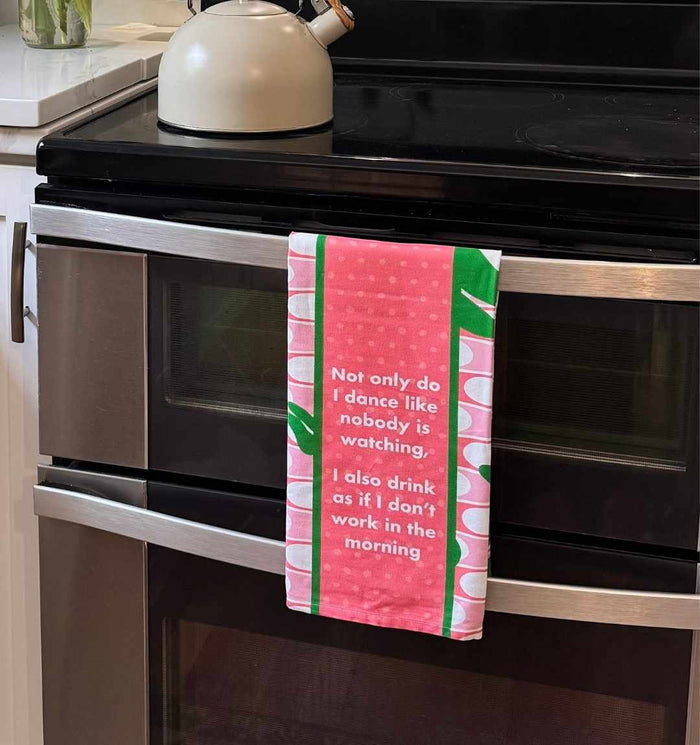 The Drinks On Me Don’t Work in Morning Dish Towel, featuring pink and green colors and a funny saying, hangs on an oven door with a white kettle on the stove above.