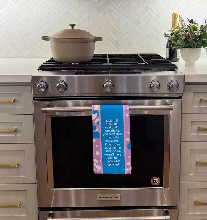 A modern stainless steel KitchenAid stove features a beige Dutch oven on top. On the oven door hangs the "Drunk Me Dish Towel" by Drinks On Me. A flower arrangement sits on the right countertop above white cabinets.