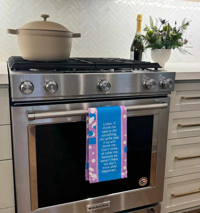 A modern kitchen stove features the Drunk Me Dish Towel by Drinks On Me hanging on the oven door, with a beige pot, champagne bottle, and flower arrangement nearby—an ideal housewarming gift for those who love kitchen humor.