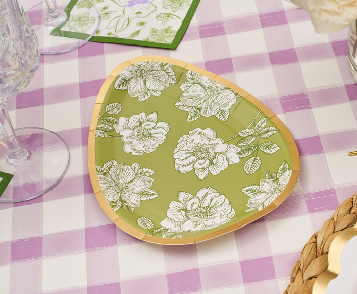 The Sophistiplate Easter Toile Assorted Die Cut Salad Plates (8pk) feature green, egg-shaped designs with white florals and a gold rim, shown atop a pink and white checkered tablecloth with glassware, napkins, and a woven placemat.