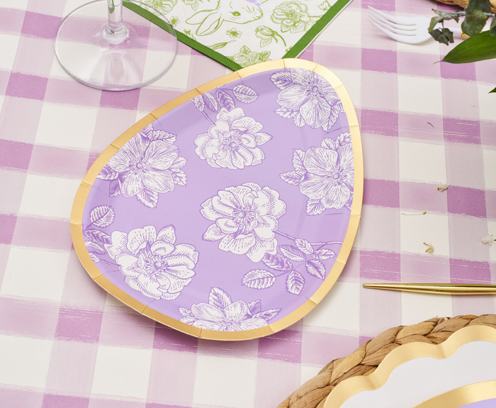 A Sophistiplate Easter Toile Assorted Die Cut Salad Plate, lavender and egg-shaped with white floral designs and a gold border, sits on a pink and white checkered tablecloth with a wine glass, napkin, cutlery, and a woven placemat nearby.