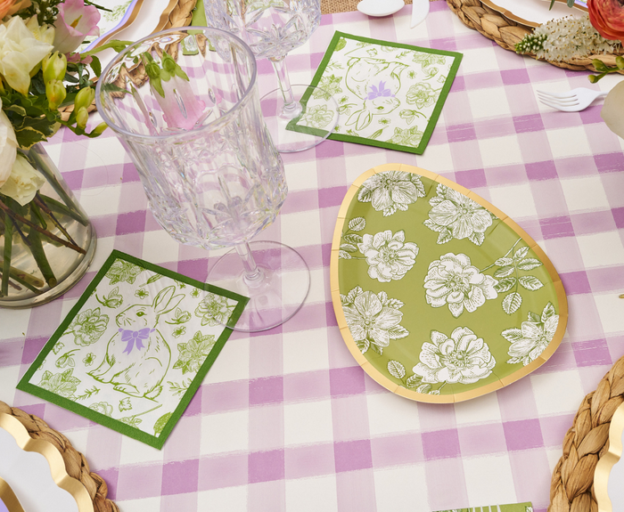 A table set with a pink and white checkered cloth, crystal glasses, floral plates, and Sophistiplate's Easter Toile Cocktail Napkins (26pk) featuring bunnies and flowers, plus a flower bouquet for charming Easter decor.