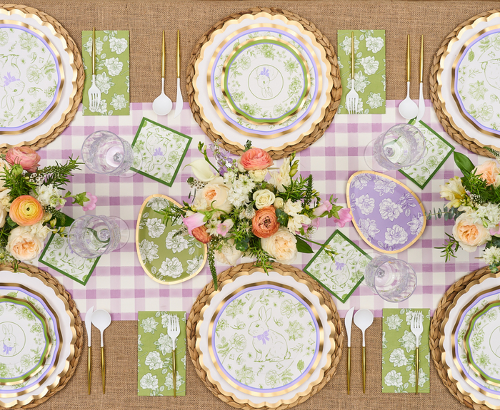 A spring-themed table with floral arrangements, playful bunny plates, green floral napkins, gold flatware, and Sophistiplate’s Easter Toile Dinner Plates (8pk) on a pink gingham runner over a burlap tablecloth.