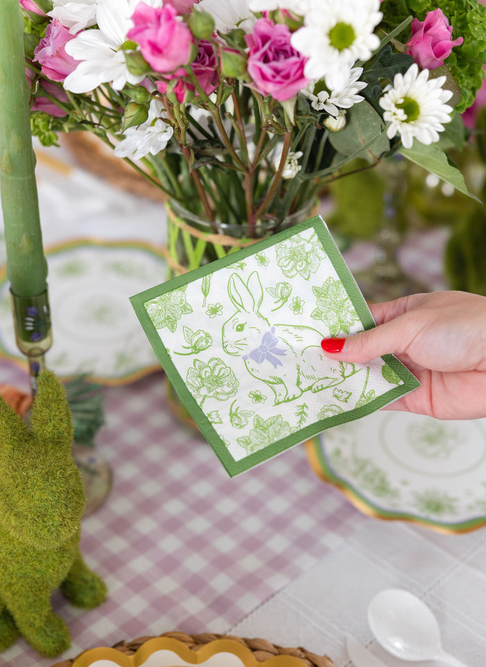 A hand with red nail polish holds a green and white bunny napkin in front of a floral centerpiece and the Sophistiplate LLC "Ultimate Easter Tablescape for 16: Easter Toile," featuring designer plates and a green moss bunny.
