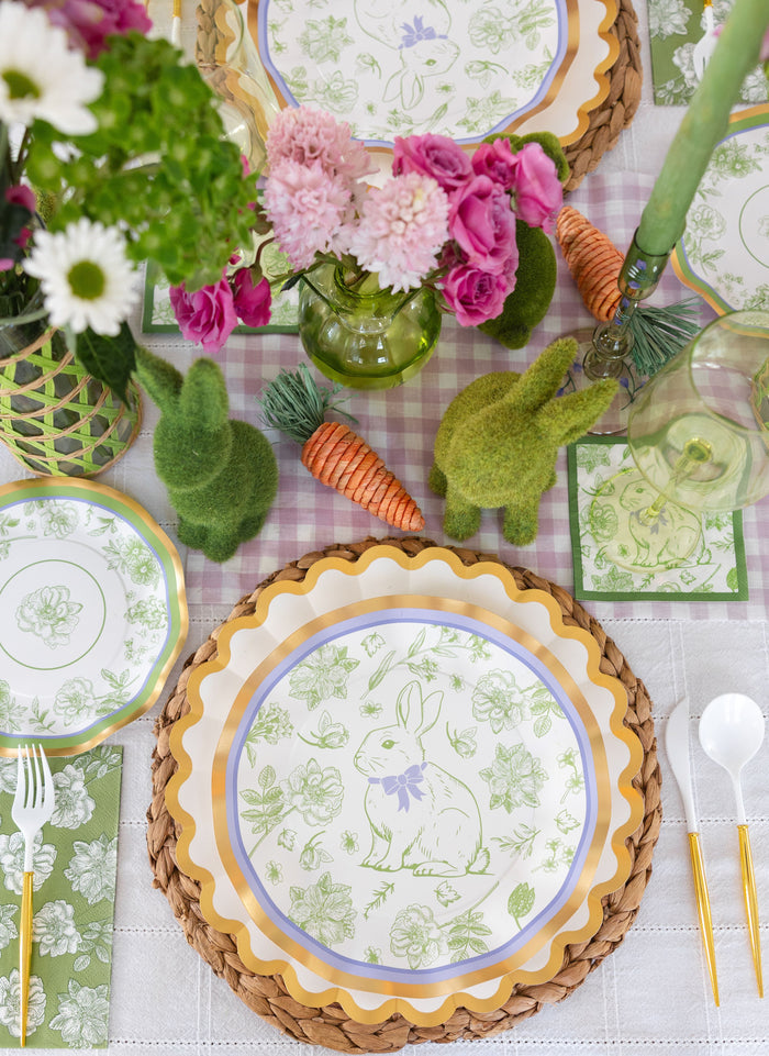The Ultimate Easter Tablescape for 16: Easter Toile by Sophistiplate LLC includes designer bunny and floral paper plates with scalloped edges, woven placemats, faux moss bunnies, carrot accents, vases of pink and white flowers, and gold-accented cutlery.