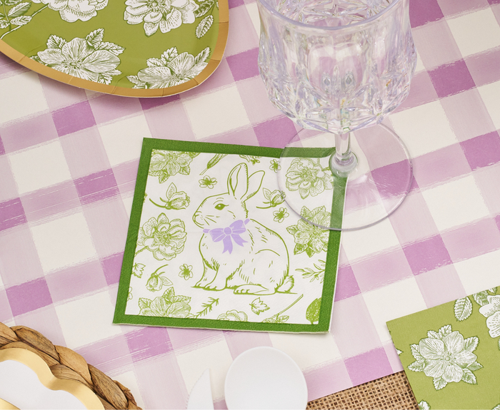A close-up of a table with a pink and white checkered tablecloth, a crystal glass, and Sophistiplate Easter Toile Cocktail Napkins/26pk featuring green and purple rabbit and floral designs.