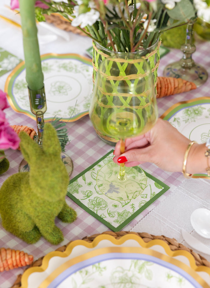 A hand with red nail polish holds a green glass on a spring-themed table decorated with Sophistiplate’s Easter Toile Cocktail Napkins (26pk), floral plates, a moss bunny, faux carrots, flowers in a vase, and Easter decor on a purple checkered tablecloth.