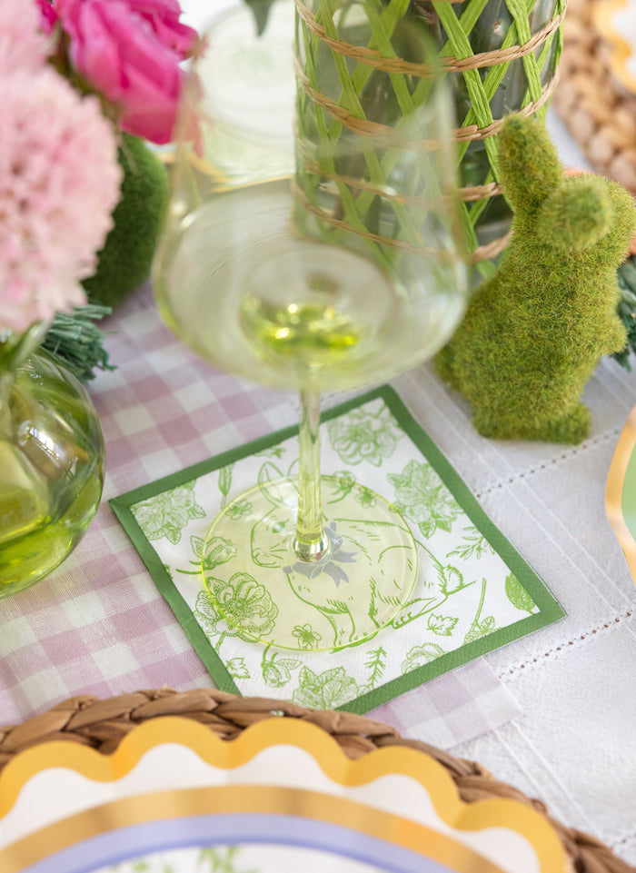 A clear wine glass rests on a Sophistiplate Easter Toile Cocktail Napkin (26pk), set atop a checkered tablecloth with pink flowers, decorative green bunny, and woven accents completing the festive Easter tablescape.