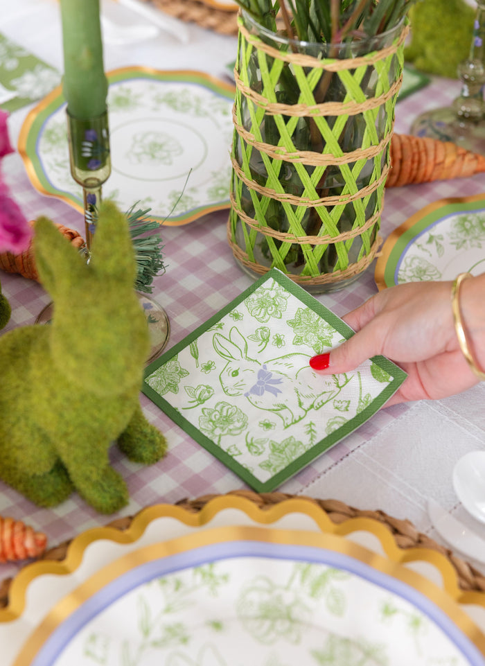 A hand holds a Sophistiplate Easter Toile Cocktail Napkin (26pk) with a green border above a festive Easter table set with bunny figurines, patterned plates, and a green candle.