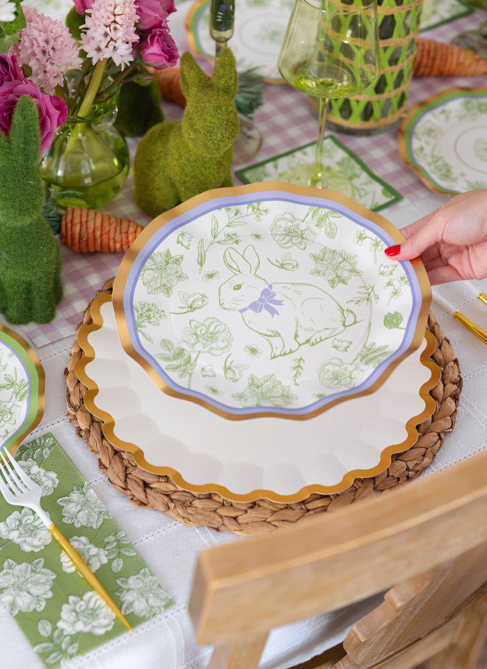 A hand places a Sophistiplate LLC Ultimate Easter Tablescape for 16: Easter Toile paper plate on a woven placemat, surrounded by green bunny figurines, flowers, and carrot decorations at an Easter tablescape.