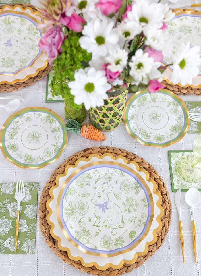Create a festive spring table with the Ultimate Easter Tablescape for 16: Easter Toile by Sophistiplate LLC, featuring bunny paper plates, green napkins, woven placemats, plastic utensils, and a white and pink floral centerpiece.
