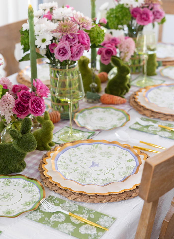 The Ultimate Easter Tablescape for 16: Easter Toile by Sophistiplate LLC includes designer floral plates, gold utensils, green napkins, pink and white flowers, moss bunnies, and green candles for a festive spring celebration.