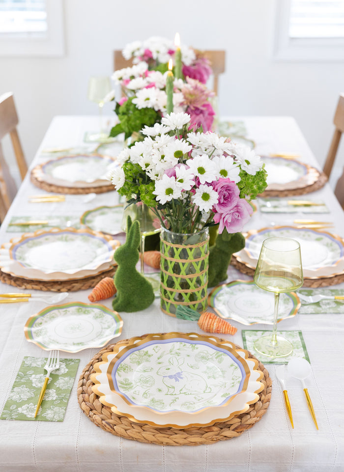 Create a spring-inspired Easter table for 16 with Sophistiplate LLC’s Ultimate Easter Tablescape: Easter Toile, featuring designer plates, wicker placemats, gold cutlery, green glasses, floral accents, bunny decor, leaf-print napkins & orange carrots.