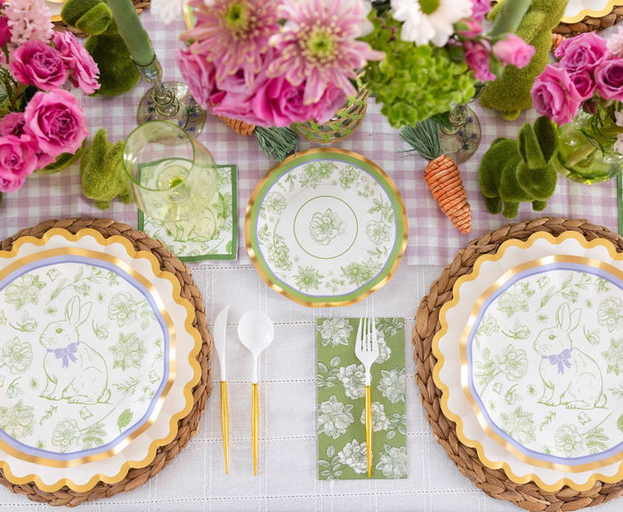 Create a festive setting with the Sophistiplate LLC Ultimate Easter Tablescape for 16: Easter Toile, featuring bunny motif plates, gold utensils, green floral napkins, wicker placemats, pink flowers, green candles, and a decorative carrot on a checkered cloth.