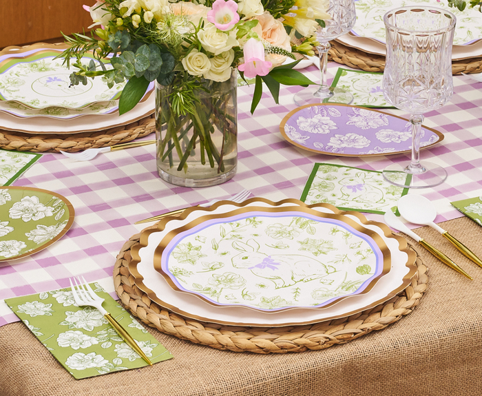 The Ultimate Easter Tablescape for 16: Easter Toile by Sophistiplate LLC features designer paper plates, gold utensils, crystal glasses, fresh flowers in a vase, a pink checkered tablecloth, woven placemats, and green and purple napkins.
