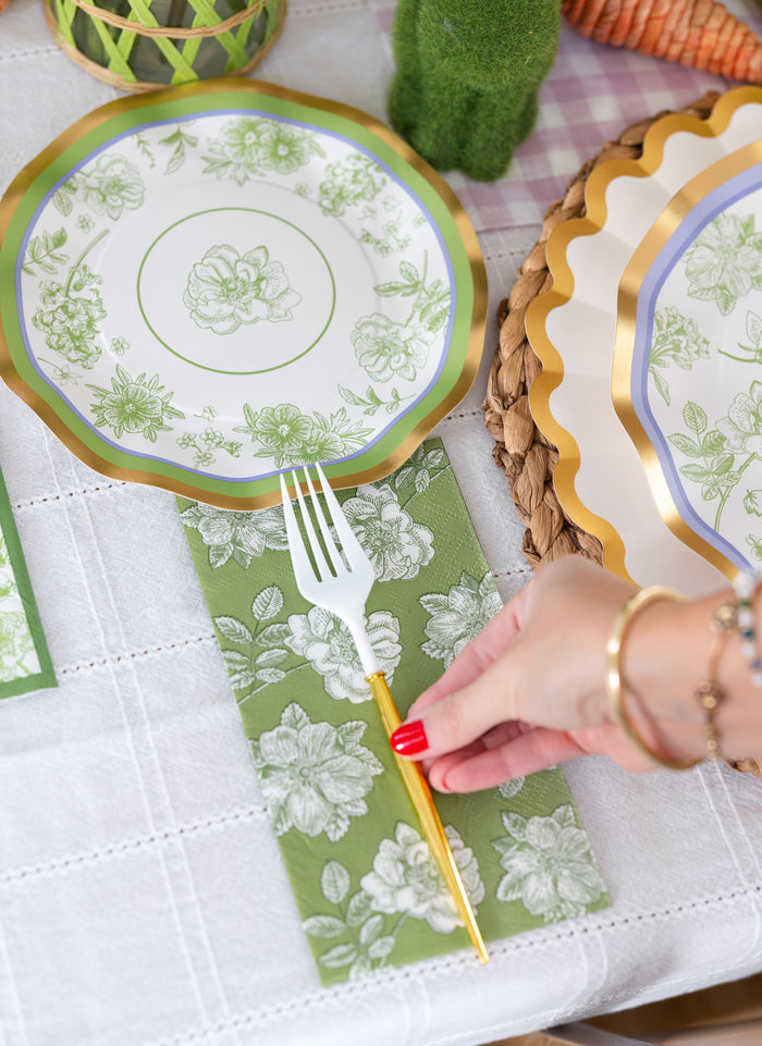 A hand with red nail polish sets a gold and white fork on Sophistiplate Easter Toile Guest Towels beside a decorative plate with a green flower pattern, all atop a white tablecloth.