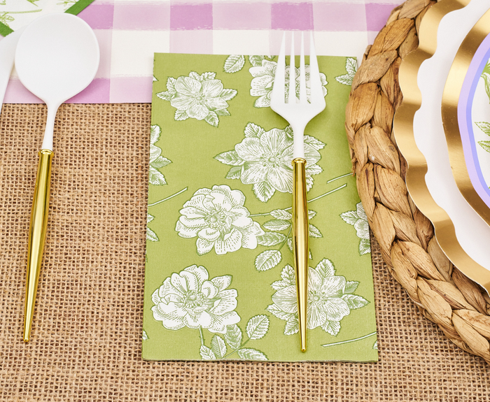 A white fork and spoon with gold handles rest on a burlap tablecloth; the fork is set atop Sophistiplate Easter Toile Guest Towels, near woven scalloped-edge plates, all on a purple checkered placemat.