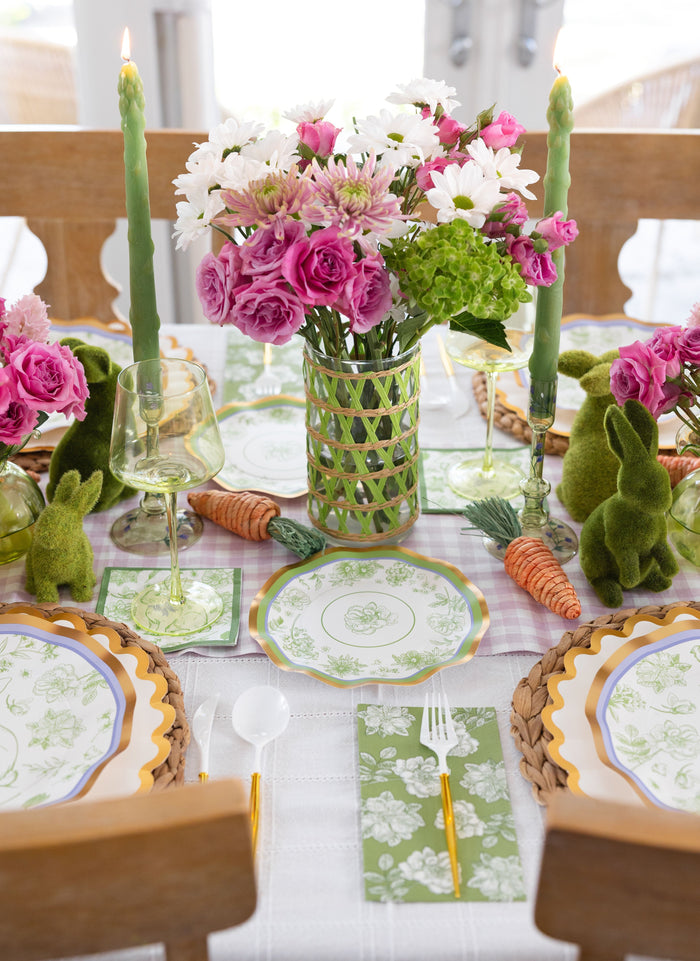 Create a spring-inspired Easter table with the Ultimate Easter Tablescape for 16: Easter Toile by Sophistiplate LLC, featuring floral paper plates, gold cutlery, green napkins, moss bunnies, carrots, candles, and a pink and white flower centerpiece.