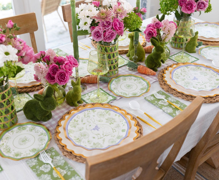 The Ultimate Easter Tablescape for 16: Easter Toile by Sophistiplate LLC features designer plates, green glasses, floral napkin-wrapped utensils, pink and white bouquets, moss bunny decor, and carrots on a white tablecloth.