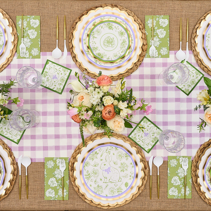 The Sophistiplate LLC Ultimate Easter Tablescape for 16: Easter Toile features bunny and floral designer plates, green floral napkins, gold utensils, clear glasses, and a pastel flower centerpiece on pink gingham over burlap.
