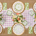 The Sophistiplate LLC Ultimate Easter Tablescape for 16: Easter Toile features bunny and floral designer plates, green floral napkins, gold utensils, clear glasses, and a pastel flower centerpiece on pink gingham over burlap.