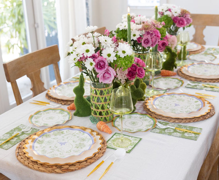 The Ultimate Easter Tablescape for 16: Easter Toile by Sophistiplate LLC includes designer paper plates, wicker chargers, green glasses, gold cutlery, and green napkins with a centerpiece of pink and white flowers, faux moss bunnies, and decorative carrots.