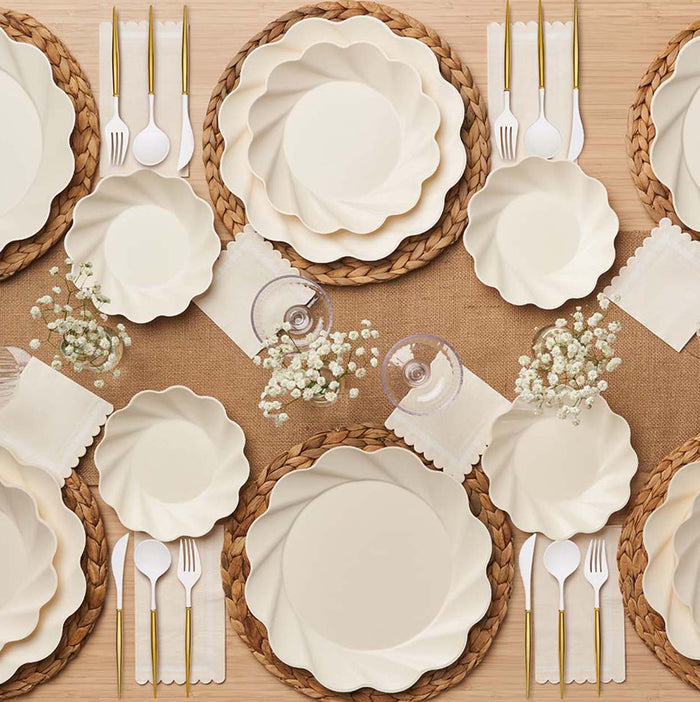 A top view of Sophistiplate's Eco-Friendly Table Setting- Cream showcases cream-hued, scalloped compostable plates, gold and white cutlery, glasses, white napkins, and baby’s breath on woven placemats with a burlap runner.