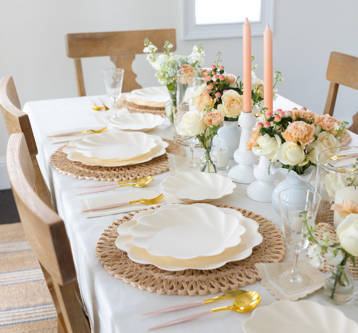 A dining table set for a meal with Sophistiplate LLC's Eco Cream & Blush Cutlery Bundle, eco-friendly plates, woven placemats, pastel pink candles, and floral centerpieces. Four wooden chairs surround the table.